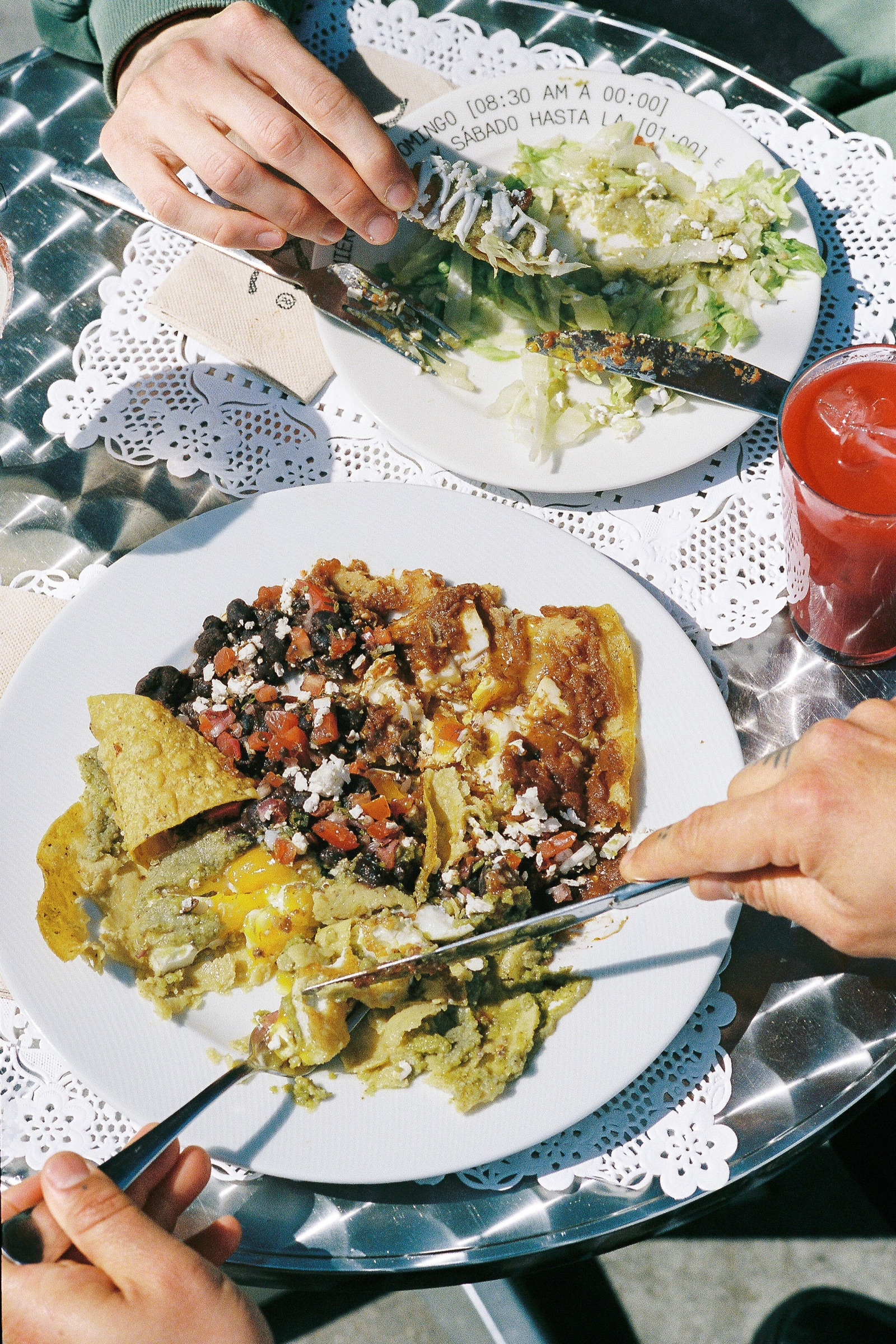Plato de enchiladas divorciadas con manos tatuadas cortando con cuchillo y tenedor sobre mesa con doily de papel y vaso de agua de jamaica.