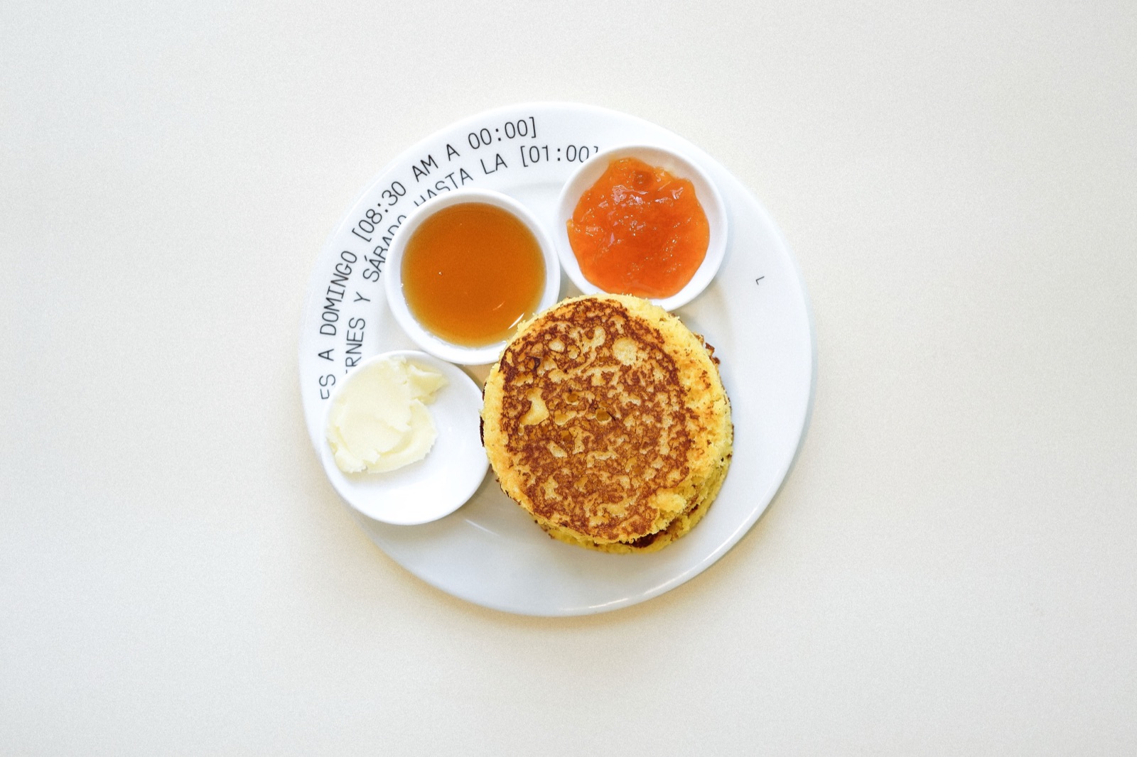 Pancakes esponjosas de maíz apiladas con miel, mermelada de durazno y mantequilla en cazuelitas blancas.
