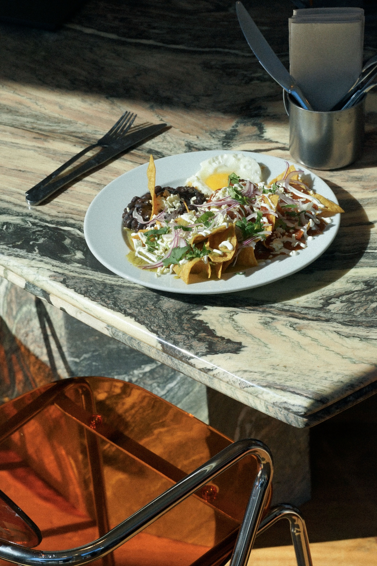 Plato de chilaquiles sobre mesa de mármol con vetas, silla ámbar y cubertería al fondo.